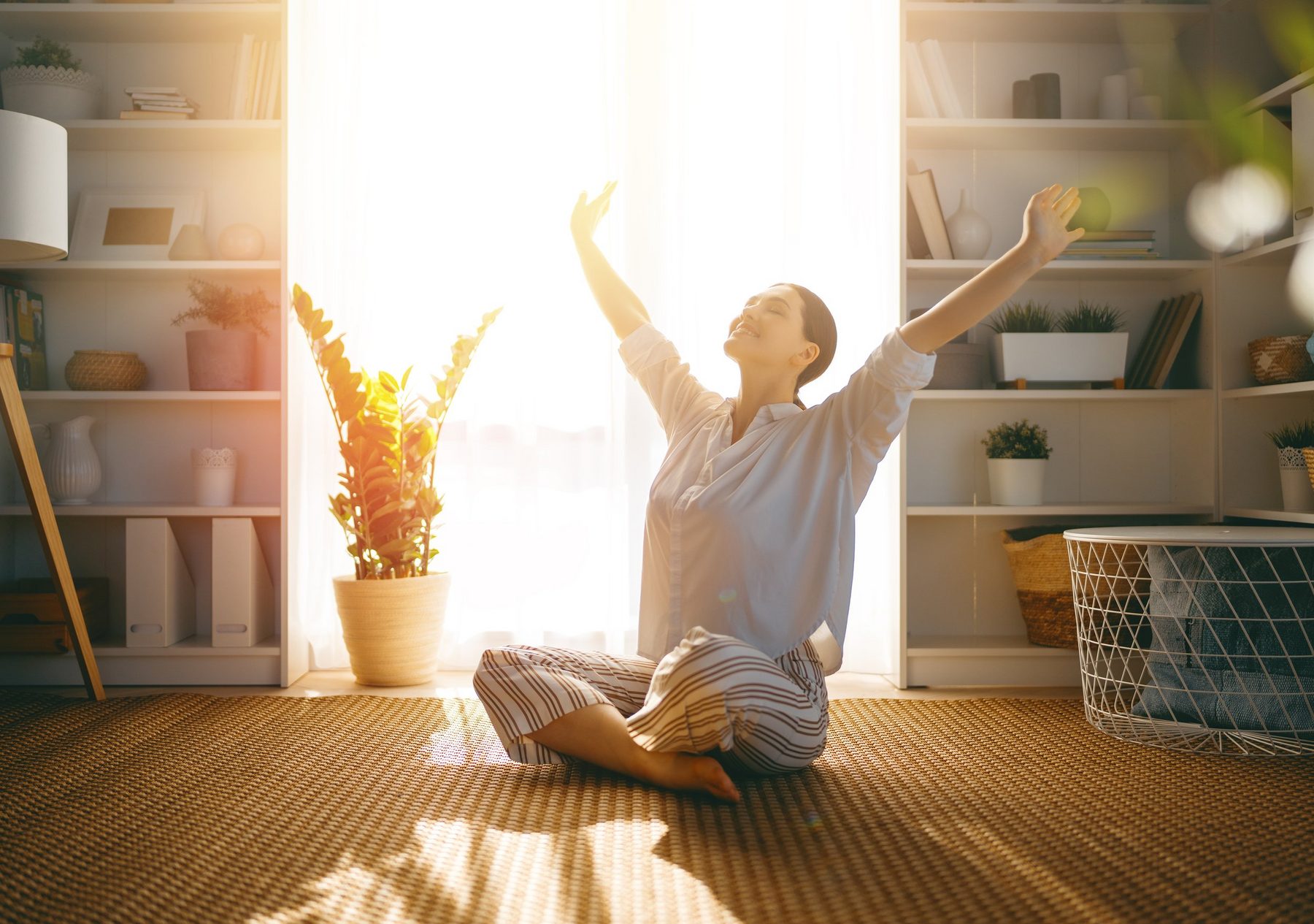 A woman stretching toward the morning light at home, arms raised, eyes closed, smiling.